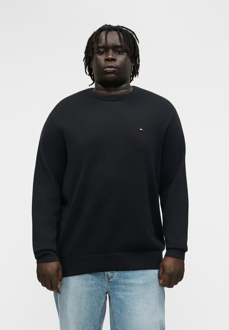 Νεαρός άνδρας με σκούρο δέρμα και ντραντλοκς φοράει μαύρο πουλόβερ με μακριά μανίκια και ανοιχτό μπλε τζιν, στέκεται μπροστά σε απλό φωτεινό φόντο.