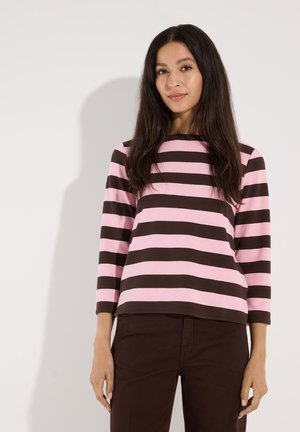 Frau mit langen dunklen Haaren, die ein rosa und braun horizontal gestreiftes Langarmshirt und dunkelbraune Hosen trägt, steht vor einem schlichten Hintergrund.