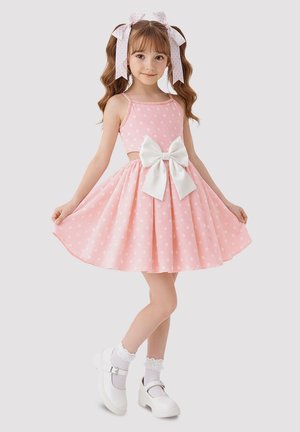Junges Mädchen trägt ein rosa Kleid mit weißen Punkten und einer weißen Schleife, passende Haarbänder, weiße Socken und weiße Mary-Jane-Schuhe und steht auf einem Bein.