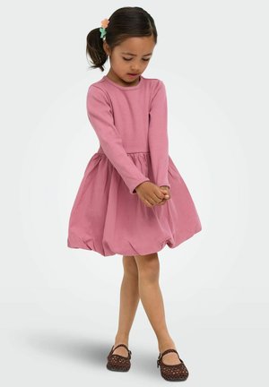 Girl with brown ponytail and flower clip wearing long-sleeve pink dress and brown woven shoes, standing with hands clasped, looking down.