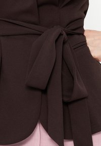 Κοντινό πλάνο σκούρου καφέ ενδύματος δεμένου στη μέση με μια φαρδιά υφασμάτινη ζώνη πάνω από ανοιχτό ροζ παντελόνια και ένα χέρι που ακουμπά στη μέση.