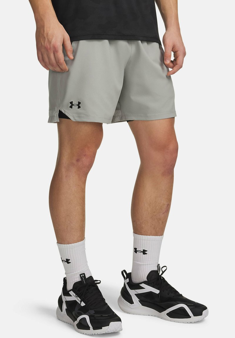 Mann trägt hellgraue Sportshorts, weiße Sneakersocken mit Logos und schwarz-weiße Sportschuhe, steht mit locker an den Seiten hängenden Händen.