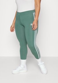 Zielone legginsy o dopasowanym kroju, z trzema białymi paskami po bokach i logo Adidas na udzie. Gładka tekstura materiału.