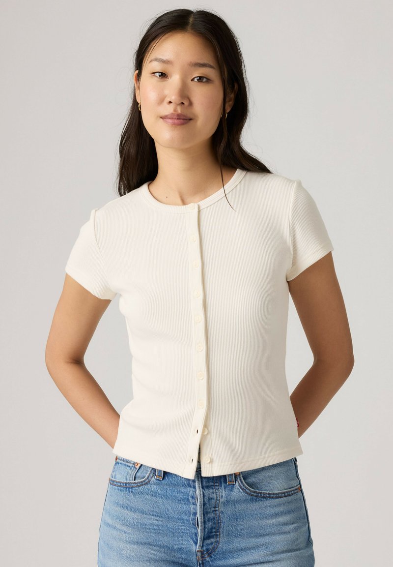 Jeune femme aux longs cheveux foncés portant une chemise blanche à manches courtes boutonnée et un jean bleu, debout les mains derrière le dos.