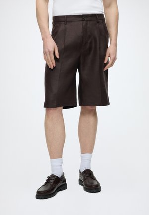 Man draagt donkerbruine op maat gemaakte knielange shorts, witte sokken, donkerbruine leren veterschoenen en een wit overhemd dat in de broek is gestopt.