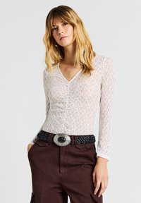 Top bianco in pizzo a maniche lunghe con scollo a V, caratterizzato da un motivo floreale; abbinato a pantaloni marroni a vita alta e una cintura nera intrecciata con fibbia argentata.