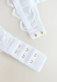 Bijele čipkaste naramenice s valovitim rubovima imaju zlatne metalne detalje i prilagodljive kuke za sigurno prianjanje.