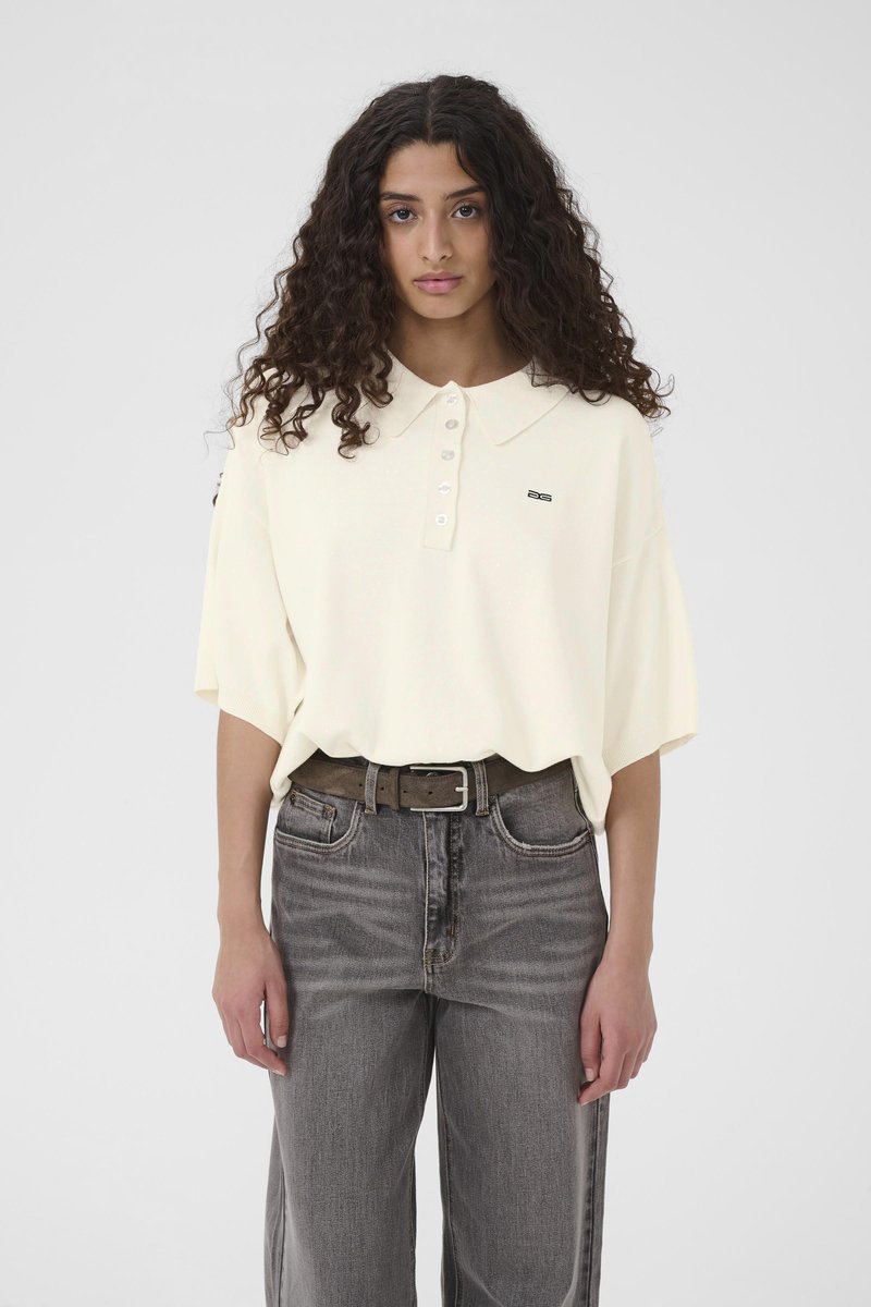 Sieviete ar cirtainiem matiem, ģērbusies brīvā balta īss piedurkņu polo krekliņā, ielocītā augsta vidukļa izbalējušās pelēkās džinsa biksēs ar brūnu jostu.