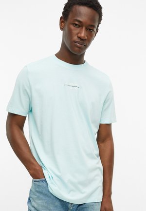 Junger Mann trägt ein hellblaues T-Shirt mit Grafik und blaue Jeans und steht mit einer Hand in der Tasche vor weißem Hintergrund.
