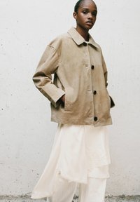 Giacca scamosciata beige con colletto classico, chiusura a bottoni e tasche laterali. Abbinata a una gonna bianca a più strati con dettagli in pizzo.