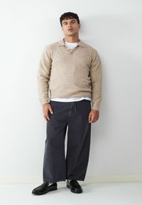 Beiger Strickpullover mit Kragen, über einem weißen T-Shirt getragen, kombiniert mit dunklen, weiten Hosen und schwarzen Loafers. Einfaches Design, strukturierter Stoff.