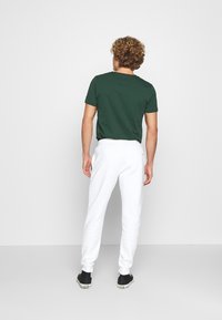 Man med lockigt hår iklädd en mörkgrön t-shirt, vita träningsoverallsbyxor och svarta högskodda sneakers, stående vänd mot en enkel vit vägg.