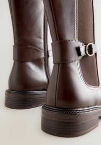 Bottes en cuir marron foncé, montant jusqu'au genou, avec une finition lisse, des panneaux latéraux élastiques et une sangle avec une boucle décorative. Talon empilé bas.