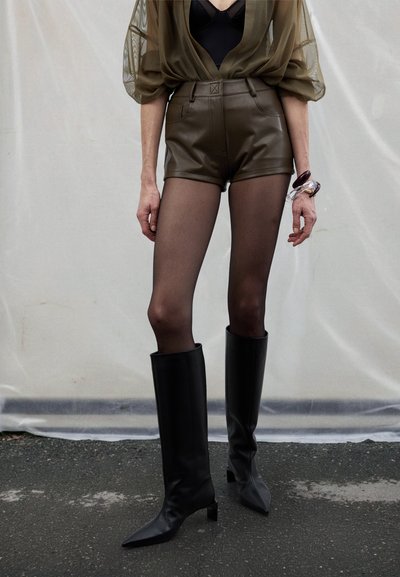 Blusa verde oliva trasparente, body nero, pantaloncini in pelle marrone a vita alta, collant neri trasparenti e stivali neri al ginocchio con punta affilata.