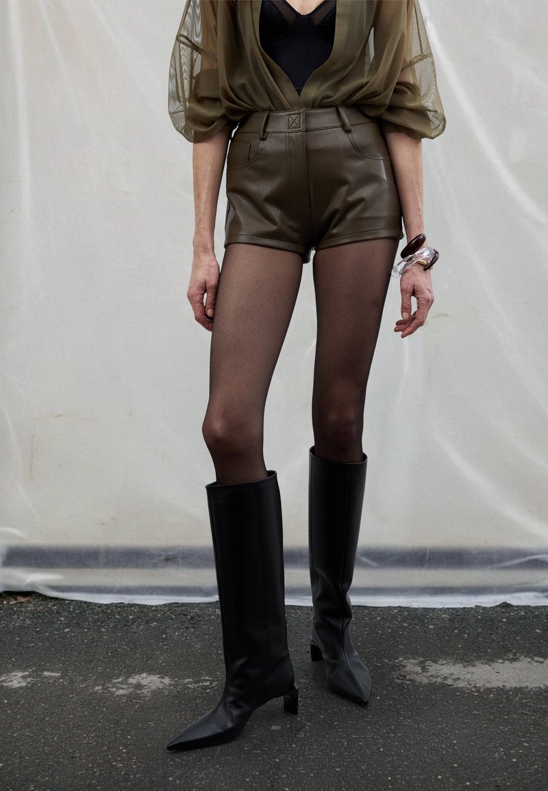 Olivazöld áttetsző blúz, fekete body, magas derekú barna bőr sort, fekete áttetsző harisnya és térdig érő fekete hegyesorrú csizma.