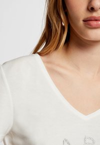 White V-neck t-shirt with short sleeves; soft cotton material featuring a subtle silver graphic design on the front. Smooth texture.