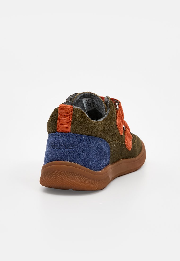 Zapatilla de ante verde con una correa naranja y un talón azul, diseño texturizado y suela de goma marrón. Cuenta con ojetes plateados para los cordones.
