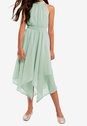Pige iført en ærmeløs, lysegrøn kjole med høj hals og asymmetrisk, lommetørklædeformet kant samt metallic ballerinaer.