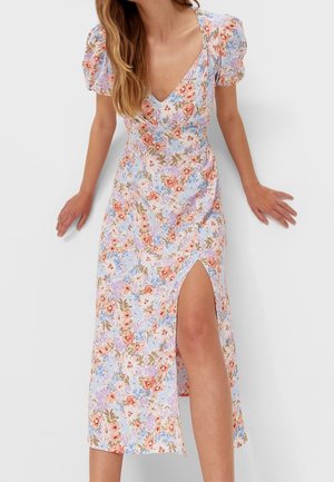 Vrouw draagt een bloemenjurk met V-hals, pofmouwen en een hoge split op de dij, staand tegen een effen lichte achtergrond.