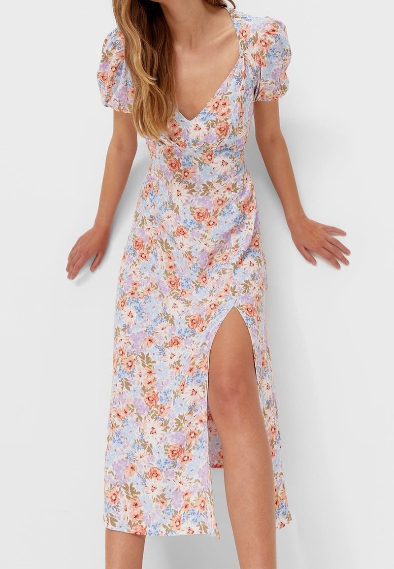 Femme portant une robe fleurie à col en V, manches bouffantes et une fente haute à la cuisse, debout devant un fond clair uni.