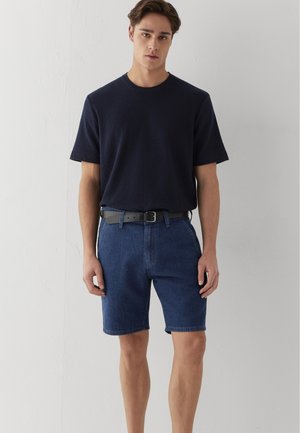 Ein junger Mann steht, trägt ein schwarzes kurzärmeliges Hemd, blaue Jeansshorts und einen schwarzen Gürtel vor einem schlichten hellen Wandhintergrund.