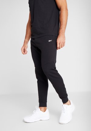 Hombre con pantalones deportivos negros de Reebok y zapatillas blancas, de pie contra un fondo gris claro liso.