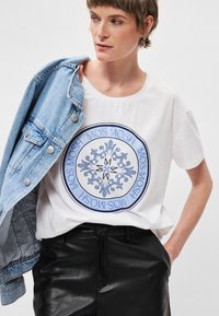 Λευκό βαμβακερό μπλουζάκι με στρογγυλό μπλε γραφικό σχέδιο που περιέχει περίπλοκα μοτίβα. Το μοντέλο φοράει ένα ανοιχτό τζιν μπουφάν πάνω από την μπλούζα.