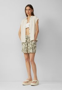 Chemise légère à boutons portée par-dessus un crop top blanc, assortie à un short à motifs verts et des sandales à plateforme beiges.