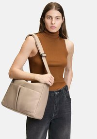 Beżowa skórzana torba na ramię z dwiema kieszeniami z przodu, miękka tekstura i regulowany pasek. Model nosi brązowy prążkowany golf i ciemne dżinsy.