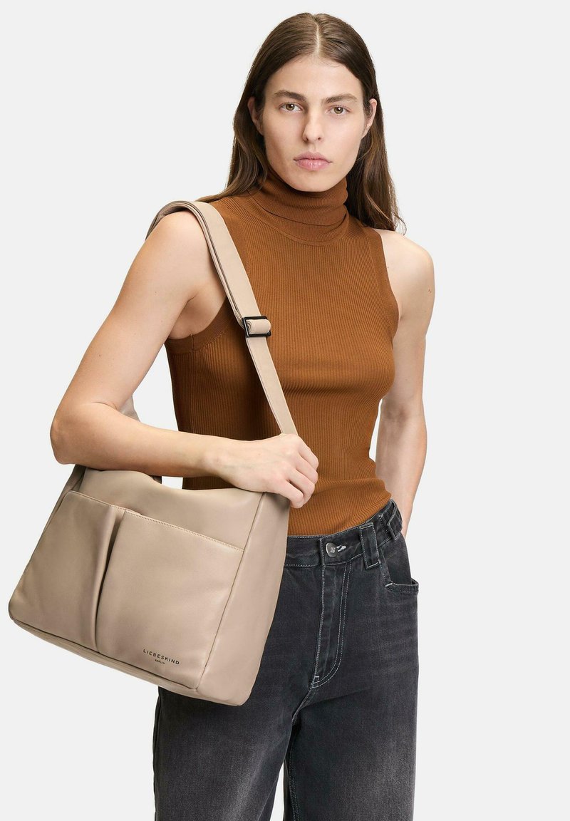 Beżowa skórzana torba na ramię z dwiema kieszeniami z przodu, miękka tekstura i regulowany pasek. Model nosi brązowy prążkowany golf i ciemne dżinsy.
