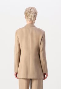 Beige strukturert blazer med glatt stoff, skarpe skuldre og en enkelt sliss bak. Tilpasset passform, minimalt med detaljer.