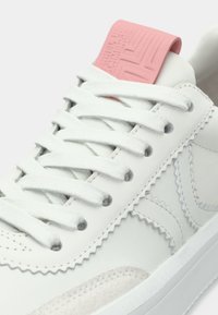 Λευκό αθλητικό παπούτσι με λευκά κορδόνια, ροζ γλώσσα, ζιγκ-ζαγκ ραφές και ανάγλυφη λεπτομέρεια στη σόλα, κοντινή λήψη.