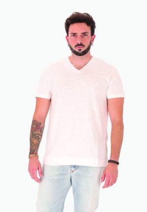 Uomo con capelli scuri corti, barba e tatuaggio sull'avambraccio, che indossa una t-shirt bianca Diesel con scollo a V e jeans azzurri chiari, in piedi contro uno sfondo bianco.
