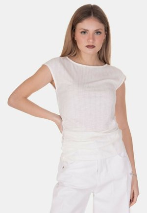 Donna con i capelli lunghi che indossa un top bianco senza maniche e pantaloni bianchi, in piedi con una mano sul fianco contro uno sfondo semplice.