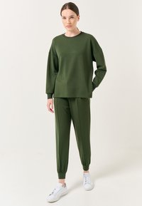 Olijfgroene sweatshirt met een relaxte pasvorm en geribbelde halslijn, gecombineerd met bijpassende tapered broek. Witte sneakers maken de outfit af.
