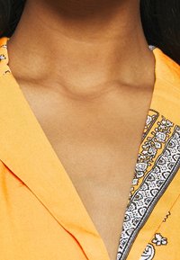 Maglietta arancione con un design a scollo a V, caratterizzata da un bordo decorativo in bianco e nero, texture liscia e vestibilità comoda.