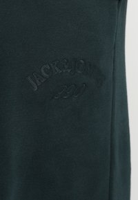 Tissu de sweatshirt vert foncé avec une texture douce. Présente un logo en relief "JACK & JONES" et "1990" dans une couleur tonale.
