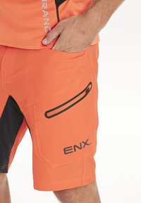 Klart orange shorts tillverkade av lättviktsmaterial. Har en svart ficka med dragkedja och texturerade detaljer. "ENX"-logotypen på sidan.