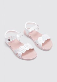 Sandalias blancas con un diseño de tira ondulada, que presentan una suela rosa y un acento metálico en forma de corazón en la tira. Textura suave, diseño casual.