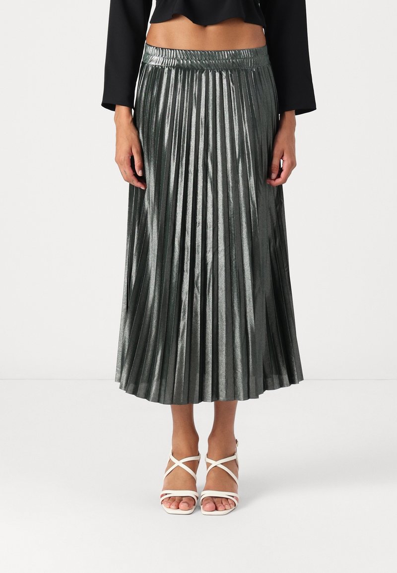 Gonna midi plissettata metallizzata verde scuro, con vita aderente e texture lucida, abbinata a décolleté bianche con cinturini sottili.