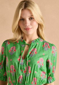 Zielona bluzka w kwiaty z falbaniastym dekoltem, ozdobiona haftowanymi czerwonymi i purpurowymi kwiatami. Krótkie rękawy z elastycznymi mankietami oraz luźny krój.