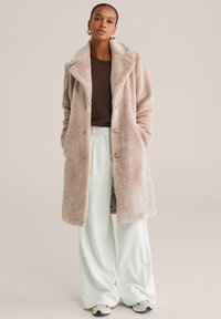 Plašči iz umetnega krzna, svetlo bež barve, z velikim ovratnikom šal stila, dvorednimi gumbi in stranskimi žepi, kombiniran z zelenkastimi širokimi hlačami.