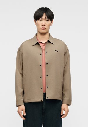 Mand iført en beige jakke med knapper over en pink polo og mørke bukser, stående foran en ensartet lys grå baggrund.