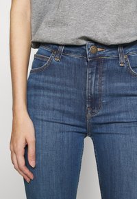 Blå denimjeans med en slim passform, med kontrasterande sömmar, fem fickor och en metallknapp i midjan.