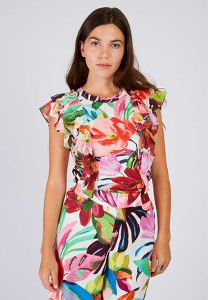 Jonge vrouw die een kleurrijke bloemen top draagt met gerimpelde mouwen en bijpassende bloemen broek met hoge taille tegen een effen achtergrond.