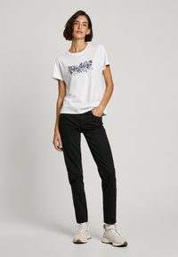 Vit kortärmad t-shirt med blommönster och texten "London", kombinerad med svarta slim-fit byxor och ljusa sportskor.
