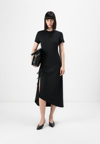 Abito nero al ginocchio con maniche corte, dettaglio arricciato ai lati e uno spacco alto. Abbinato a una borsa nera e scarpe ballerine a punta.