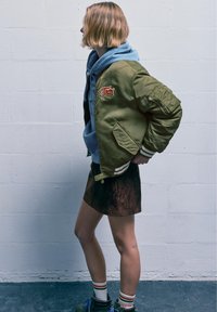 Oliivinvihreä pompierejacke, jossa on paikka, kerroksittain vaalean sinisen hupparin päällä. Ruskea mokkaminihame ja raidalliset sukat täydentävät asua.