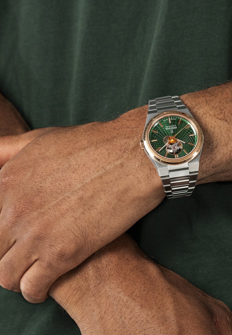 Sudraba un zelta pulkstenis ar zaļu teksturētu ciparnīcu, ar redzamu kustību un nerūsējošā tērauda aproci. Valkāts uz rokas.