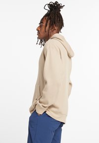 Beige ribbed hoodie with a front pocket and adjustable drawstring. The model wears navy blue pants, showcasing a relaxed fit.